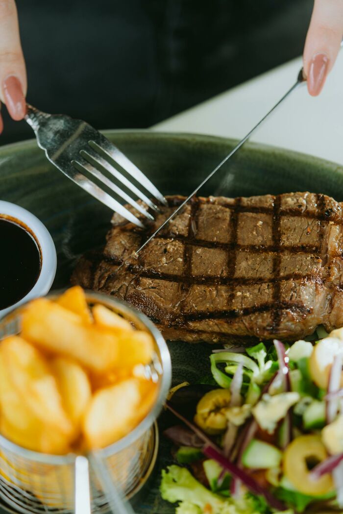 Person cutting into a grilled steak on a plate with potatoes and salad, alongside a small bowl of sauce.