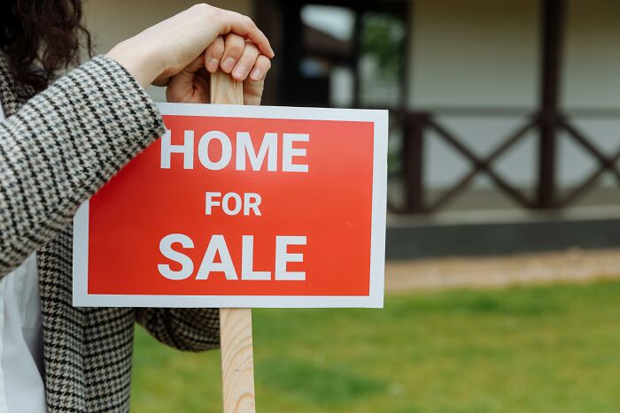 Person holding a home for sale sign, standing outside with greenery in the background.