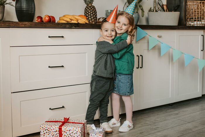 Two kids in party hats hugging, next to gifts and a kitchen counter, evoking being picked last in gym class.