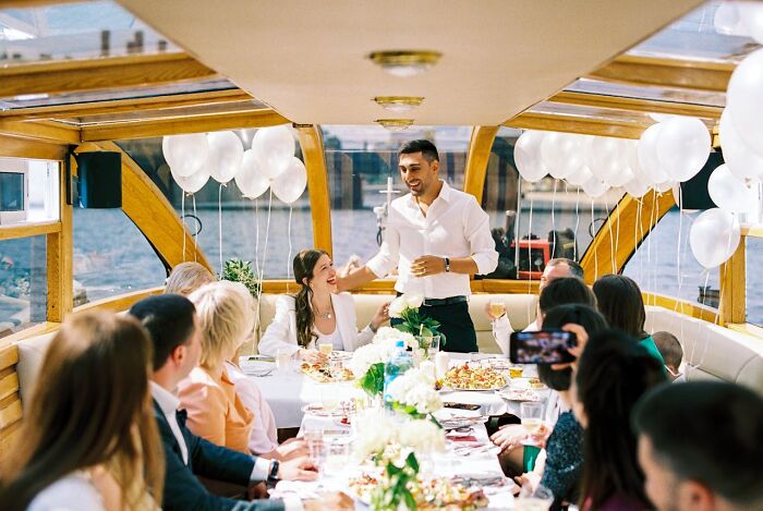 A man gives a speech at a wedding reception on a boat, surrounded by guests and white balloons.