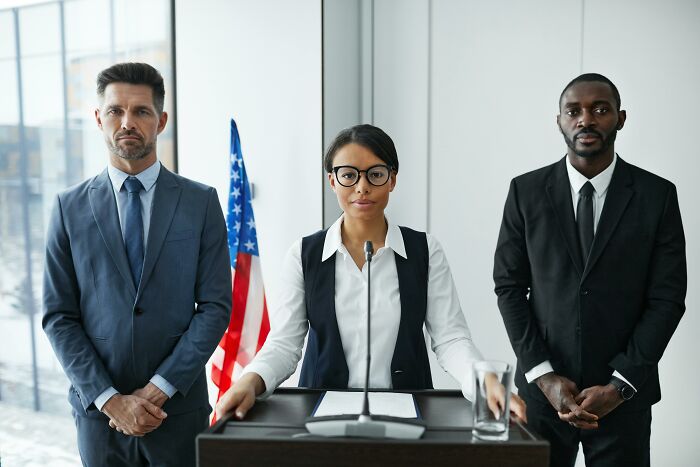 Two men in suits and a woman at a podium, with the American flag, discussing something society finds normal.