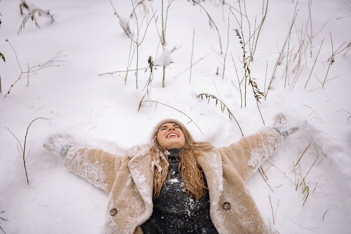 Person in a fur coat lying joyfully in the snow, surrounded by winter scenery.