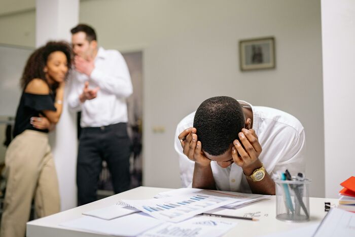 A stressed employee at a desk, while two coworkers whisper, illustrating unprofessional behavior at work.