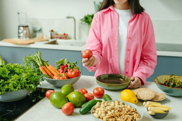 Person in a pink shirt holding a tomato in a kitchen with a variety of fresh vegetables displayed on the counter.