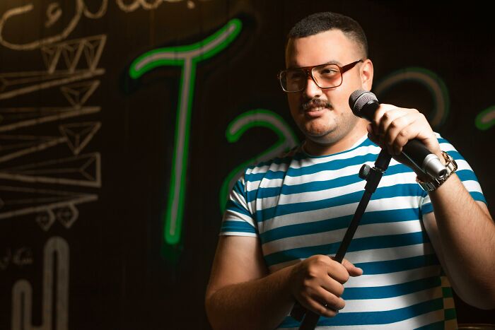 Man in glasses holding a microphone, wearing a striped shirt, standing against a dark background with neon designs, indicating intelligence.