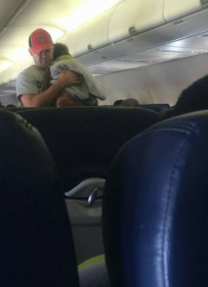 Man in red cap tenderly holding a small child wrapped in a blanket on an airplane, showing a positive and uplifting moment.