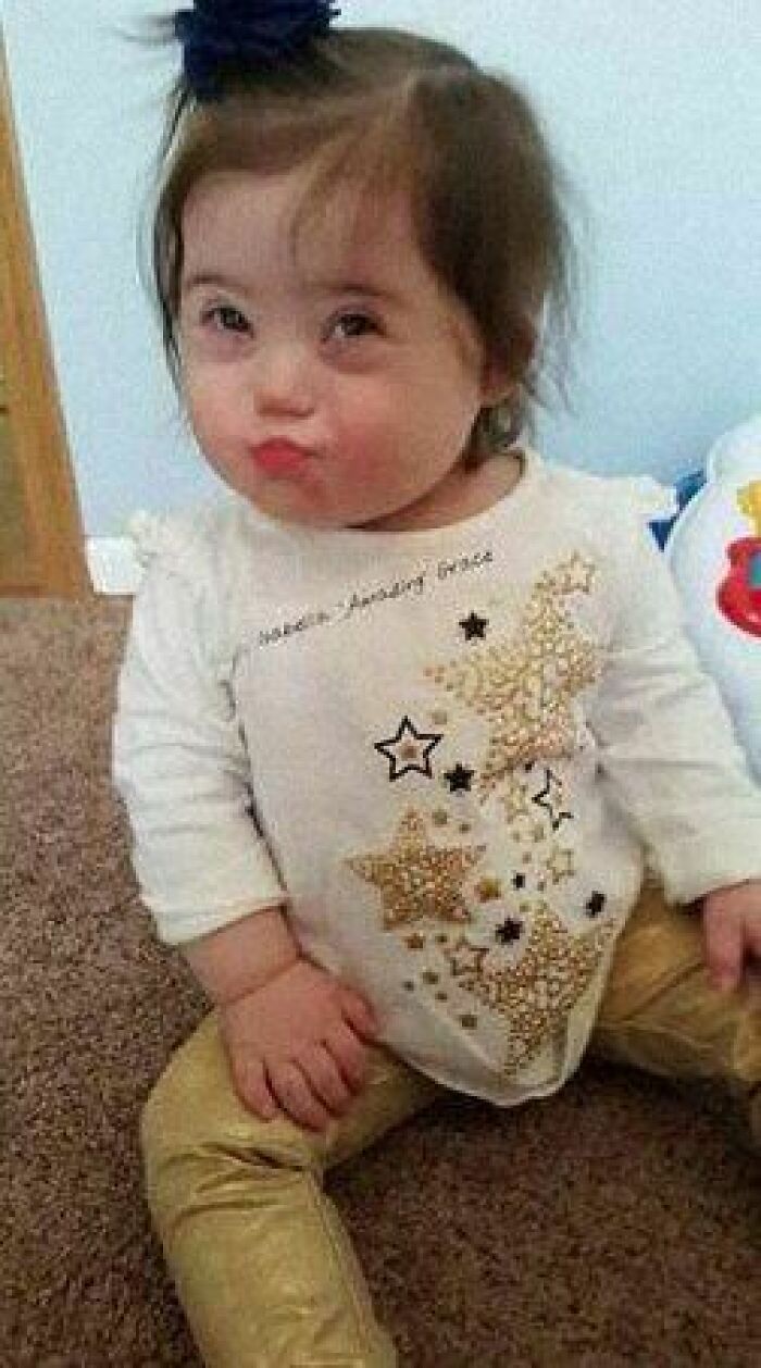 Adorable child with a playful expression, wearing a decorative shirt and gold pants, on a carpeted floor. Positive vibes only.
