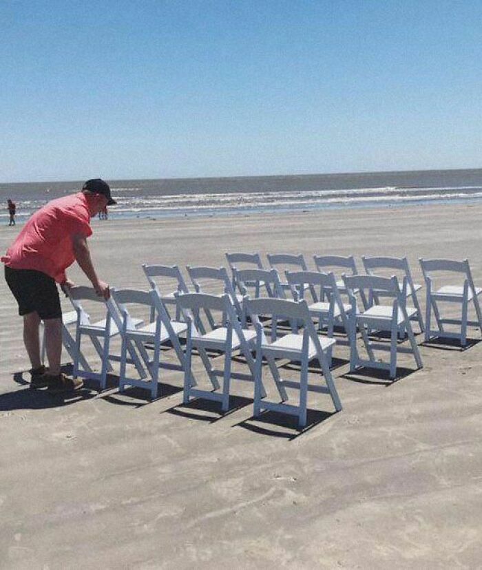 Man arranging white chairs on a sandy beach under a clear blue sky; uplifting post showcasing simple outdoor setup.