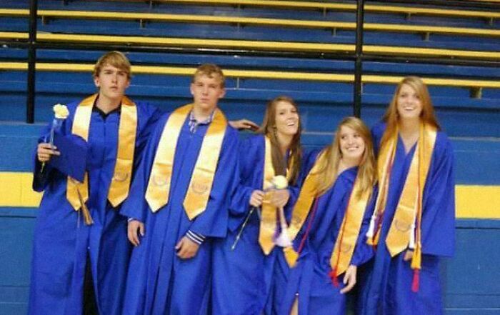 Graduating students in blue robes and gold stoles, smiling and holding flowers, sharing a positive moment.