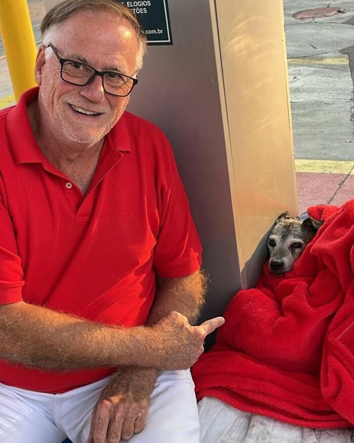 Man in red shirt smiling next to a stray dog wrapped in a red blanket at a gas station. Man in red shirt smiling next to a stray dog wrapped in a red blanket at a gas station.
