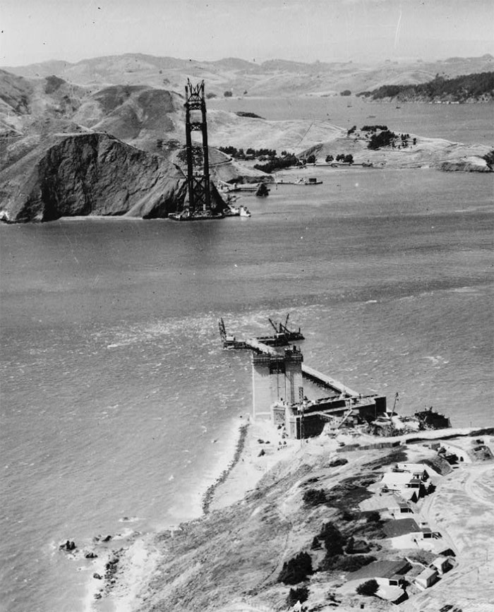 Early construction of the iconic Golden Gate Bridge, with cranes and partial structure over the water.