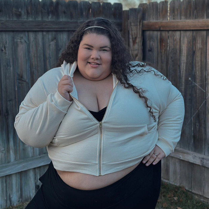 Plus-size woman confidently wearing a white hoodie in front of a wooden fence.
