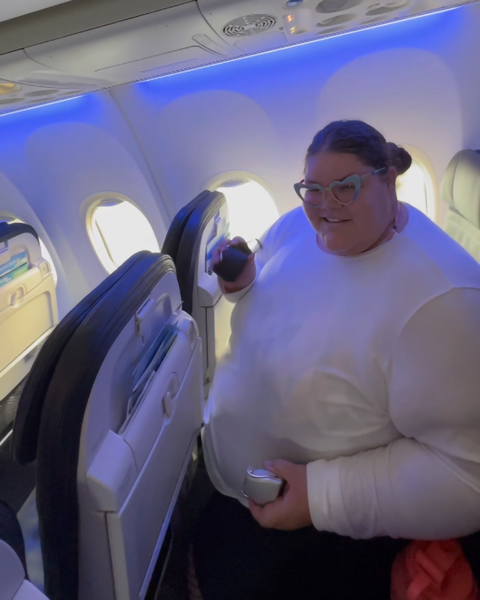 Plus-size woman in airplane seat, holding a phone, addressing airline seating policies.