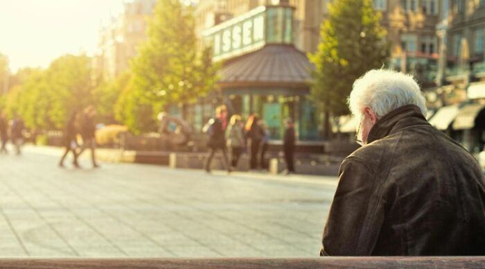 Elderly man sitting alone on a bench in a sunny plaza, facing buildings and trees, pondering life's dilemmas.