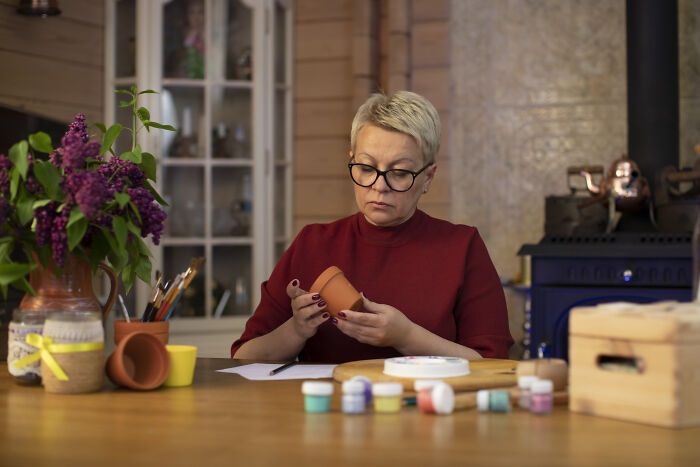 Person examining a small pot at a wooden table with paints and brushes, engaging in a hobby often underrated by many.