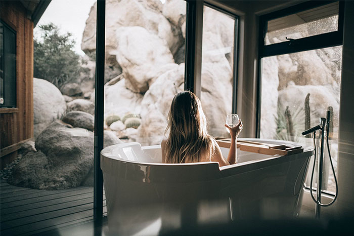 Person in a bathtub with a scenic view of rocks, holding a glass, representing a social media moment.