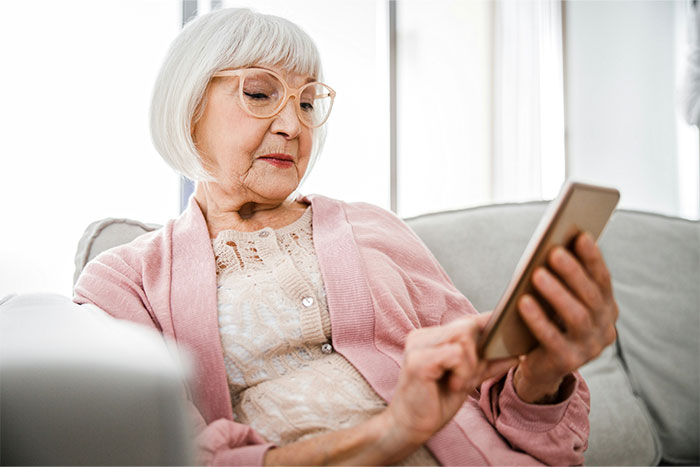 Elderly woman in pink cardigan, using a smartphone to browse social media on a cozy sofa.