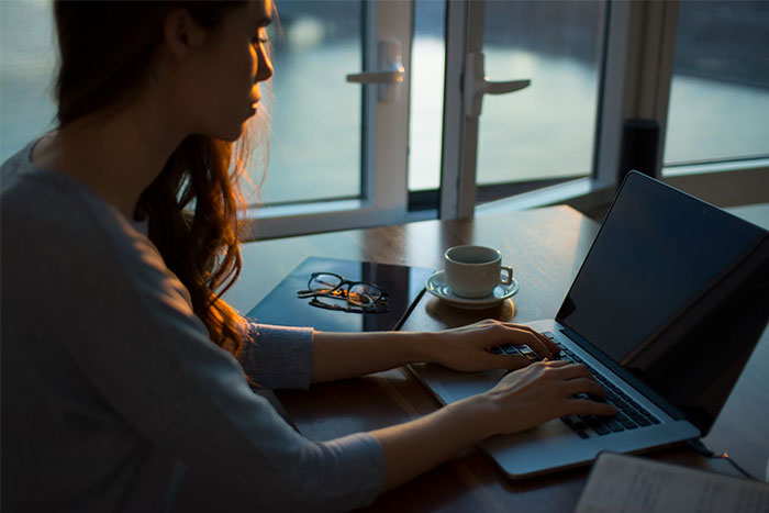 A woman typing on a laptop near a window, possibly posting to social media, with a coffee and glasses nearby.