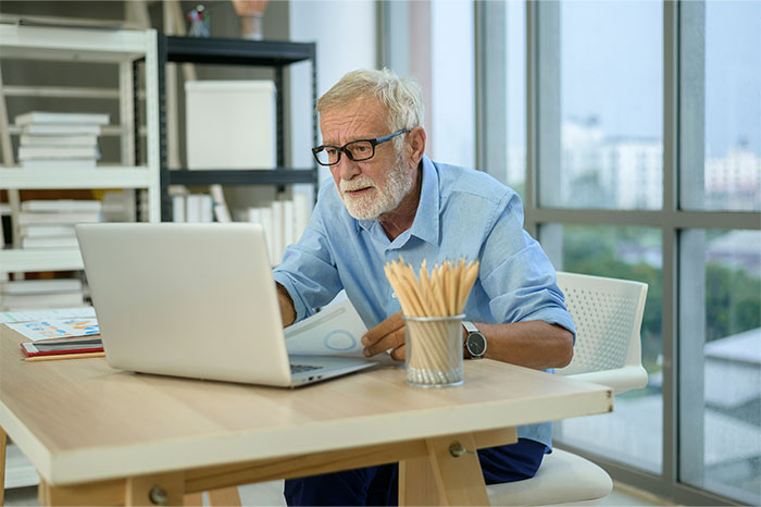 Elderly man in glasses using a laptop at a desk with bookshelves, illustrating social media content creation.