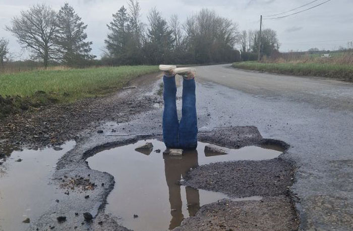 Legs sticking out of a pothole on a rural road; prank alerts officials to local hero&rsquo;s concern.