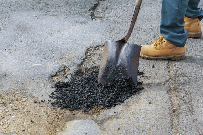Worker filling pothole with asphalt, wearing yellow boots, using a shovel.