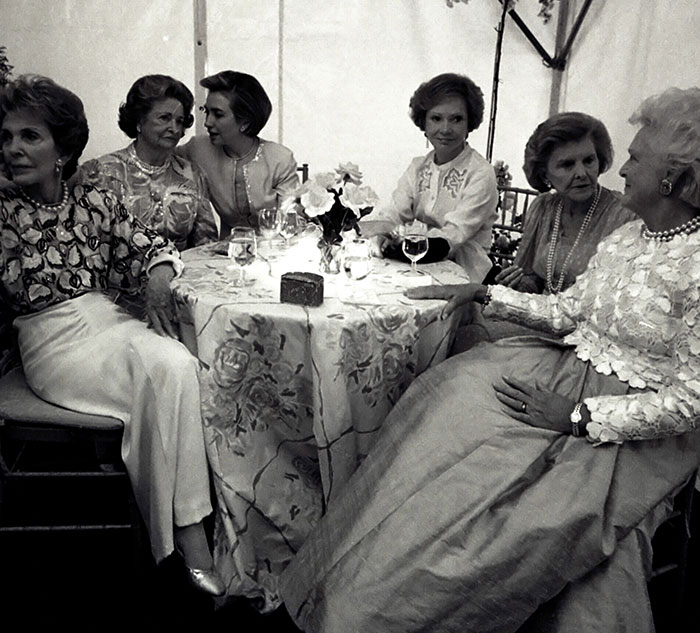 Group of women seated around a table, engaging in conversation, wearing elegant attire. Fascinating social gathering scene.