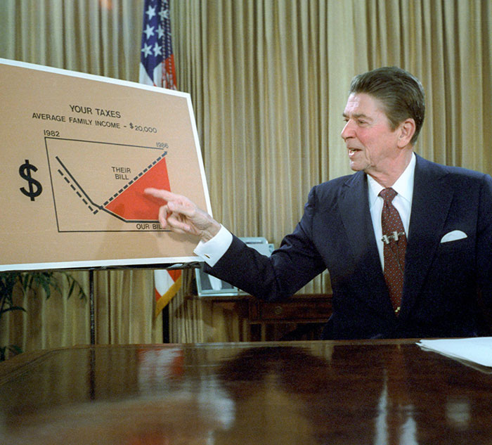 Man in a suit discussing a tax chart, pointing to data; an opportunity to learn new economics facts today.