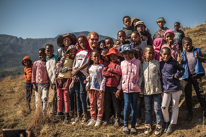 A man in a Harvard shirt surrounded by smiling children outdoors in a mountainous area, related to charity and controversy.