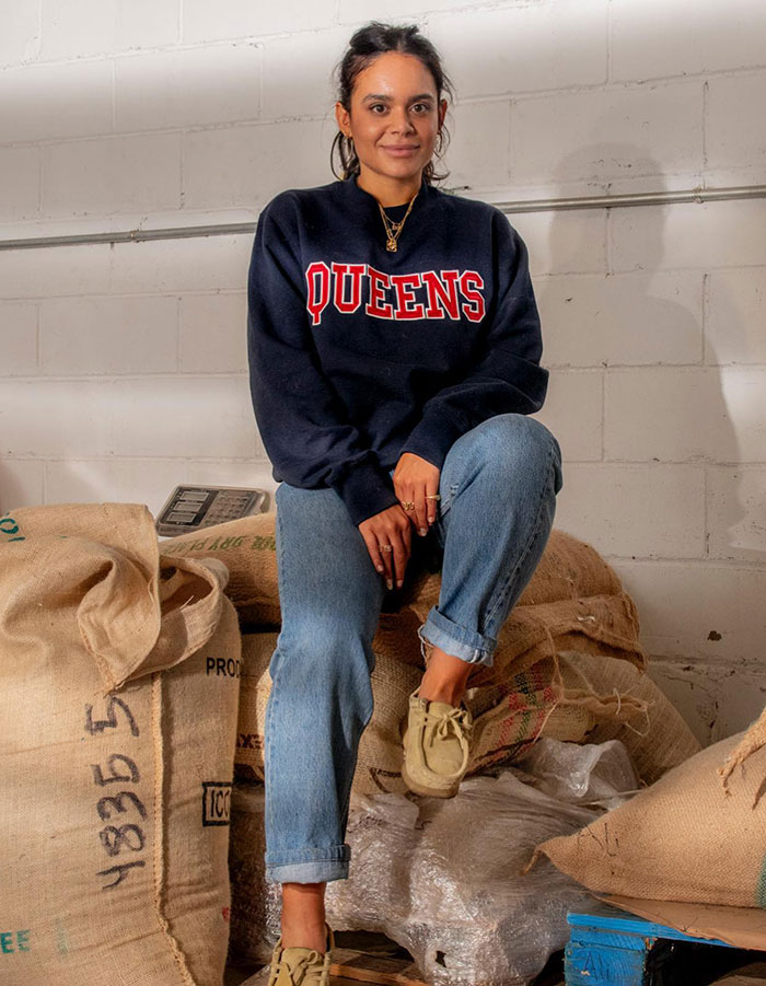 Smiling cafe owner sitting on coffee bags in NYC, wearing a "Queens" sweatshirt and jeans. Smiling cafe owner sitting on coffee bags in NYC, wearing a "Queens" sweatshirt and jeans.