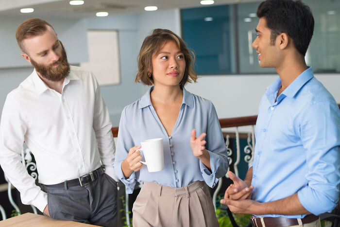 Three people engaged in conversation, one holding a mug, discussing signs of being raised in a cave in an office setting.
