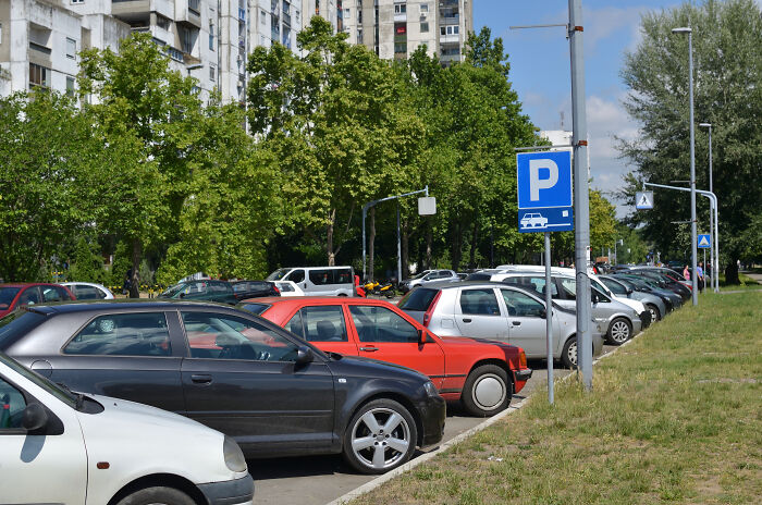 Cars parked in a row near a blue parking sign with trees and buildings in the background, highlighting urban development.