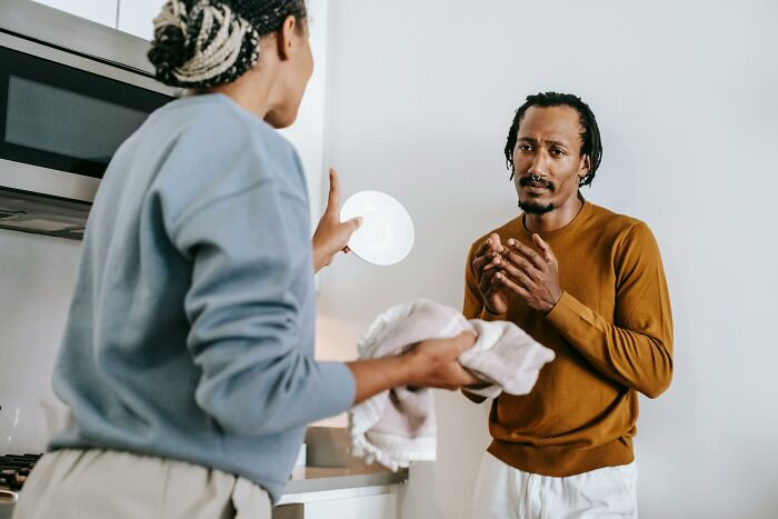 Two people in a kitchen, with one holding a dish, discussing signs someone was raised in a cave.