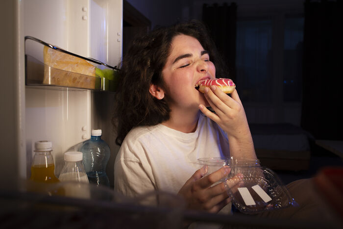 Person eating a donut in front of an open fridge, representing a telltale sign someone was raised in a cave.