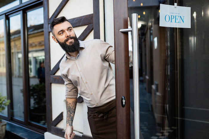 Man with a beard holding a door open, wearing a beige shirt.