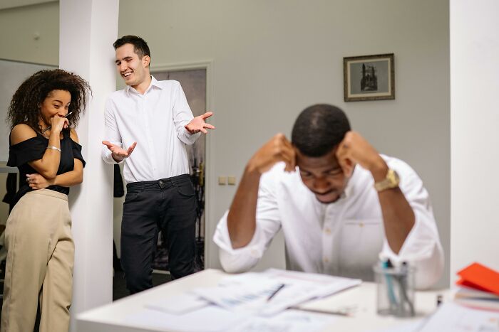 Two people laughing while a third person looks frustrated at a desk, highlighting social signs someone was raised in a cave.