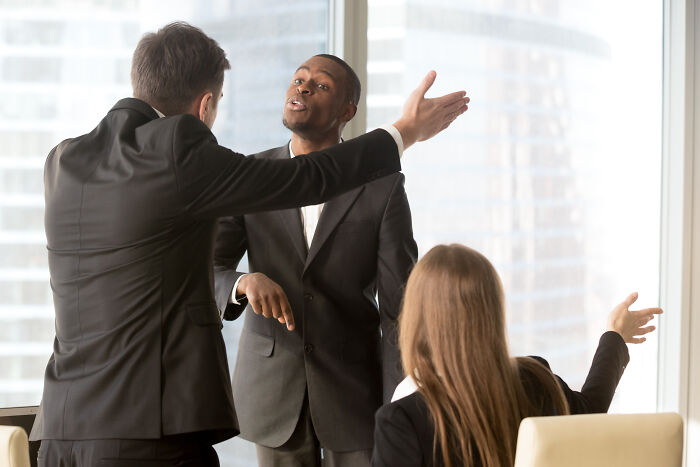 Two men in suits arguing intensely, with a woman pointing, discussing signs of being raised in a cave.