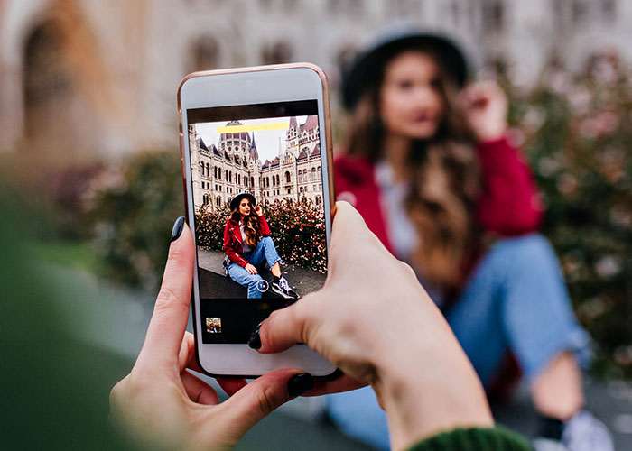 Person taking a photo of a friend posing in front of a building, illustrating friendship dynamics.