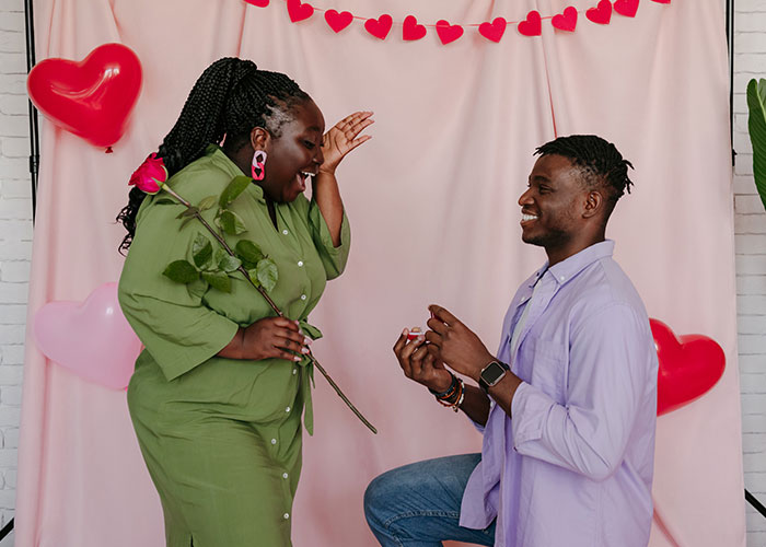 Man proposing to excited woman holding rose, with heart decorations in the background.