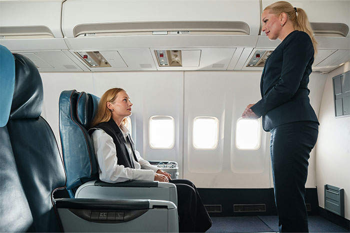 Passenger sits in airplane seat while flight attendant stands nearby discussing seat arrangements.