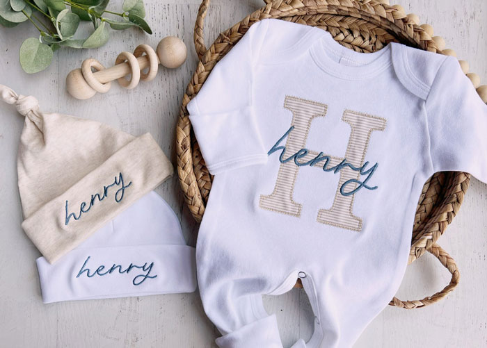 Baby clothes with "Henry" name, breaking tradition, on a woven basket with a wooden toy and greenery nearby.