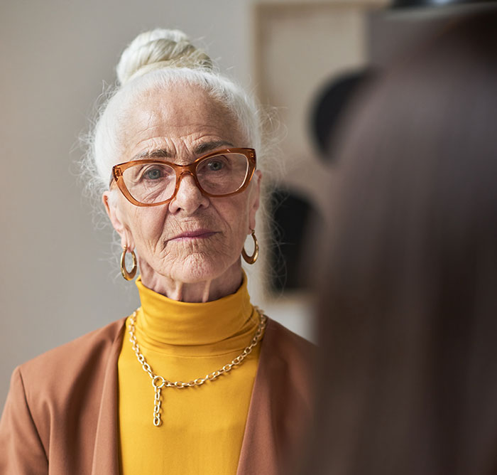 Elderly woman with glasses and white hair in a bun, wearing a mustard turtleneck and brown blazer, looking concerned. Elderly woman with glasses and white hair in a bun, wearing a mustard turtleneck and brown blazer, looking concerned.