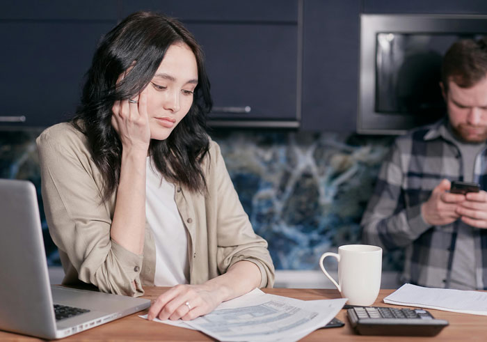 Woman concerned over college fund papers at kitchen table, man using phone in background.