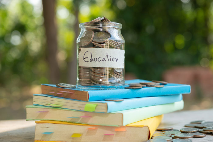 Jar labeled "Education" filled with coins on stacked books, illustrating a college fund concept.