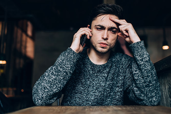 Man in a gray sweater on a phone call, looking concerned, in a dimly lit cafe, discussing U.S. tariffs and 51st state jokes.