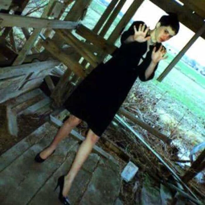 A woman in a black dress and heels, hands raised in a creepy pose inside an old wooden structure.