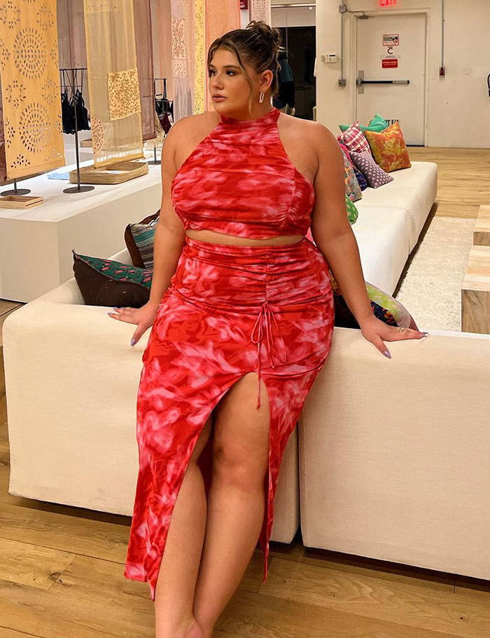 Plus-size influencer seated on couch, wearing a red patterned dress, in a stylish, modern setting.