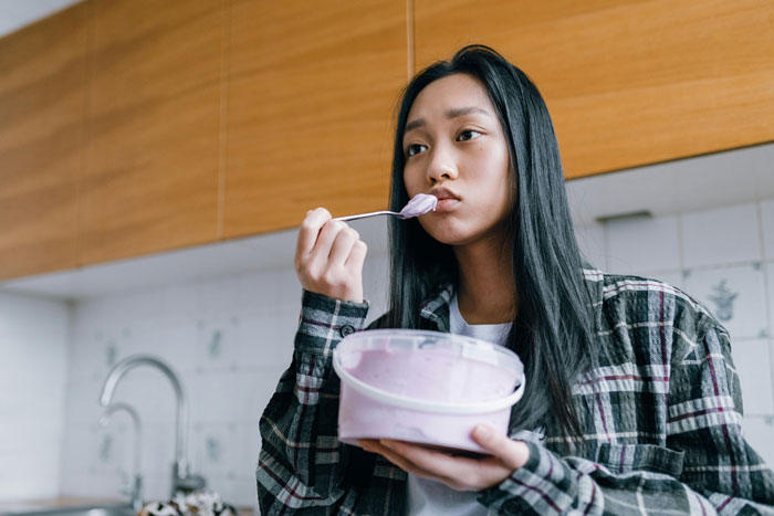 Young woman with a plaid shirt eating ice cream in a kitchen, tasting a surprise flavor.