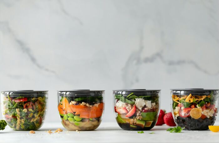 Four meal prep containers with fresh ingredients on a marble counter, illustrating frugal hacks in food preparation.