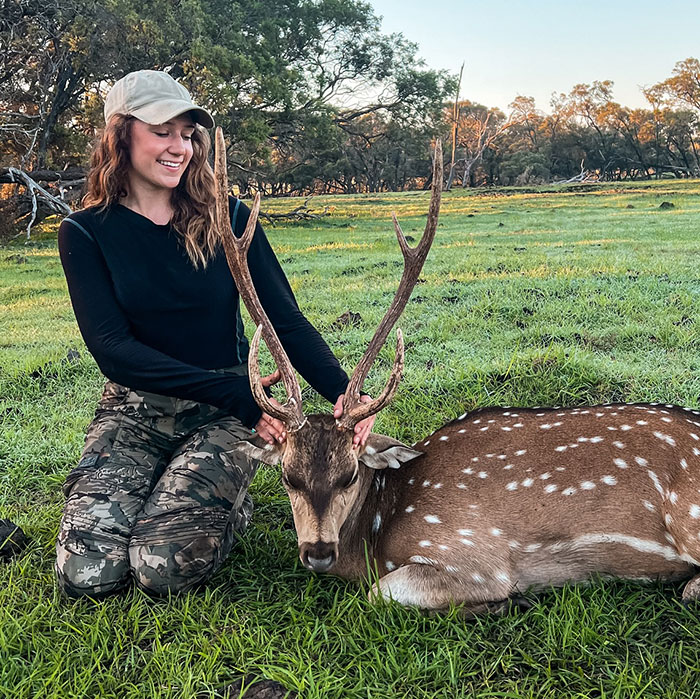 Woman in a hat and camouflage pants kneeling next to a resting deer in a grassy field. Woman in a hat and camouflage pants kneeling next to a resting deer in a grassy field.