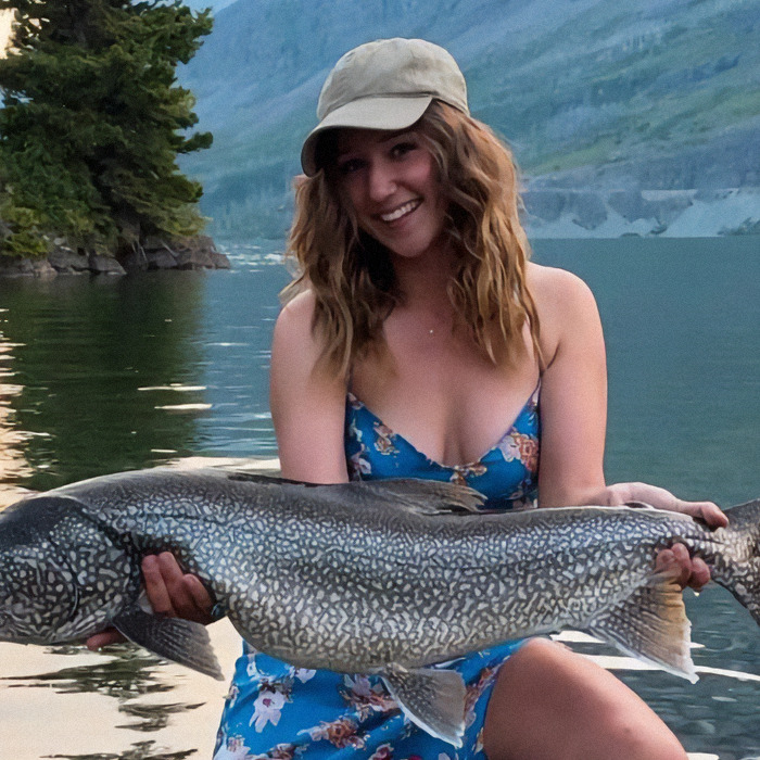 Woman in a floral dress holding a large fish by a lake, smiling; related to American tourist and baby wombat incident. Woman in a floral dress holding a large fish by a lake, smiling; related to American tourist and baby wombat incident.